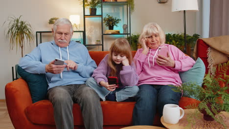 child girl granddaughter with grandparents spending leisure time using smartphones at home on sofa