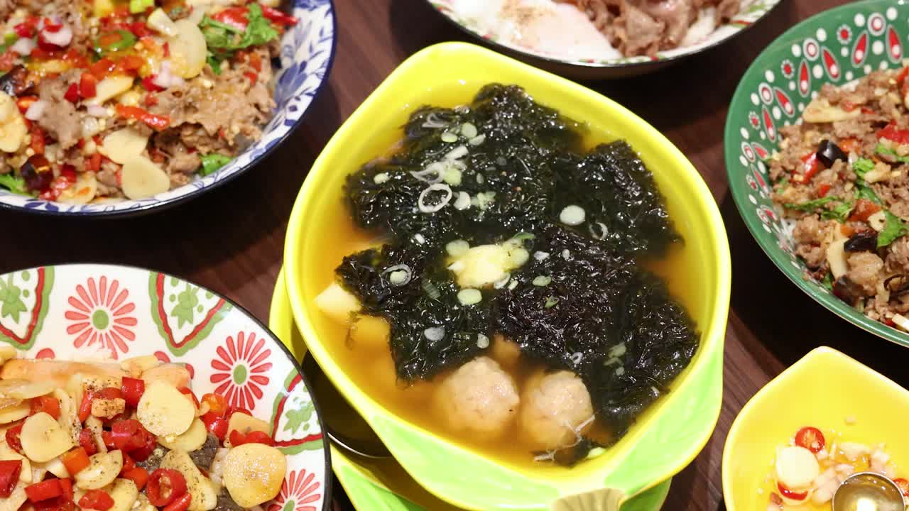 A vibrant assortment of Thai rice dishes, soups, and sides is displayed on a wooden table under bright lighting, captured in a smooth overhead camera movement