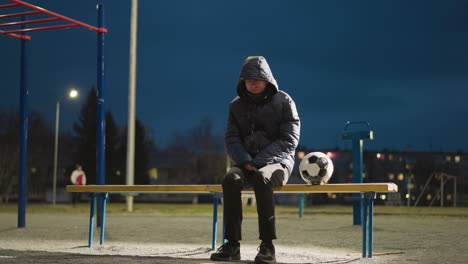 a man sits thoughtfully on a bench outdoors at night in a quiet stadium, with his hands clasped between his legs, a soccer ball by his side, with another person walks in the background