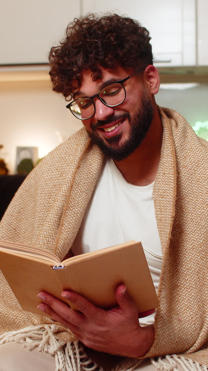 Un homme arabe portant des lunettes lit son livre préféré emmitouflé dans une couverture, lors d’une soirée tranquille, détendue et paisible.