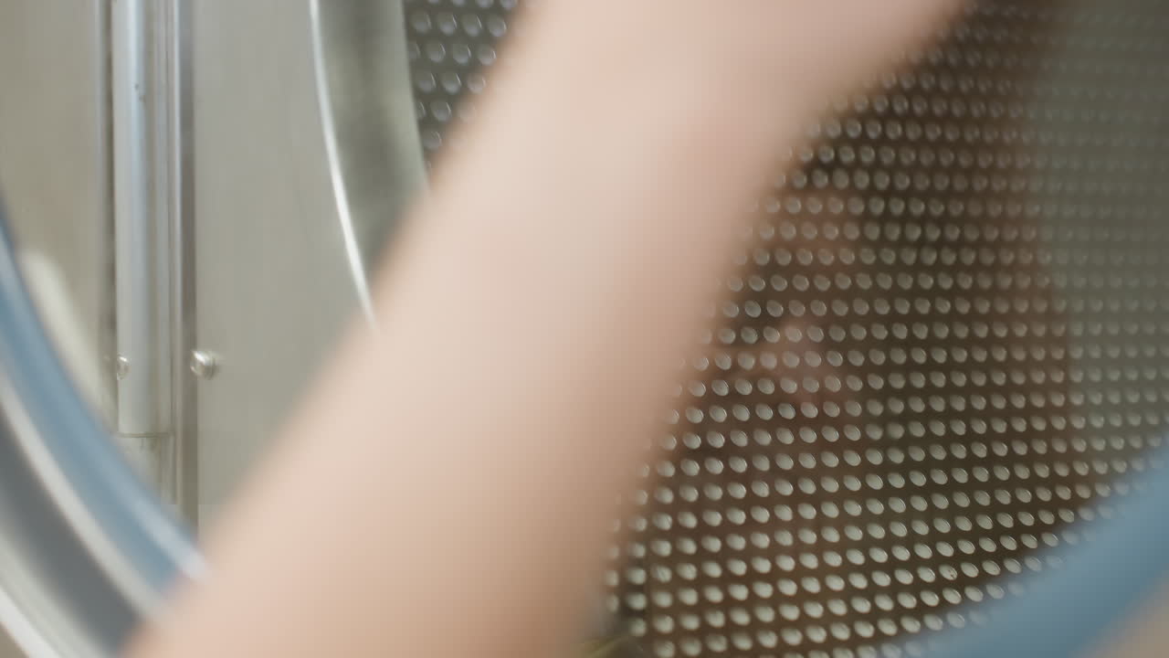 Close up hand view of laundry attendant wiping transparent surface door of washing machine with soapy towel, leaving streaks of water and reflection visible on glass while performing cleaning task