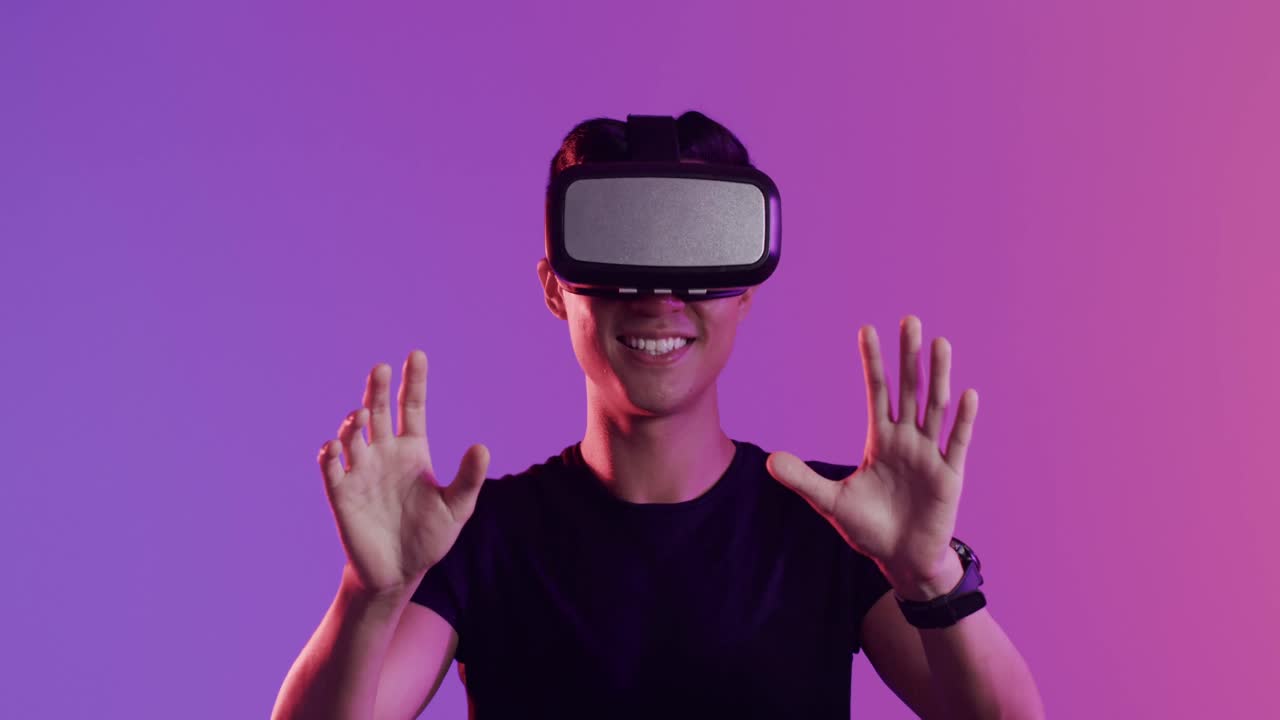 Happy asian man using vr headset and touching virtual screen on purple background, slow motion