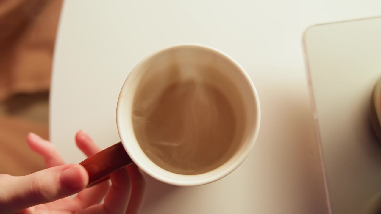 Una vista dall'alto mostra una tazza di caffè in ceramica con vapore che sale e sottili increspature sulla superficie, mentre una mano si allunga verso la tazza su un tavolo rotondo bianco, con luce calda e soffusa e un'atmosfera interna calma e accogliente.