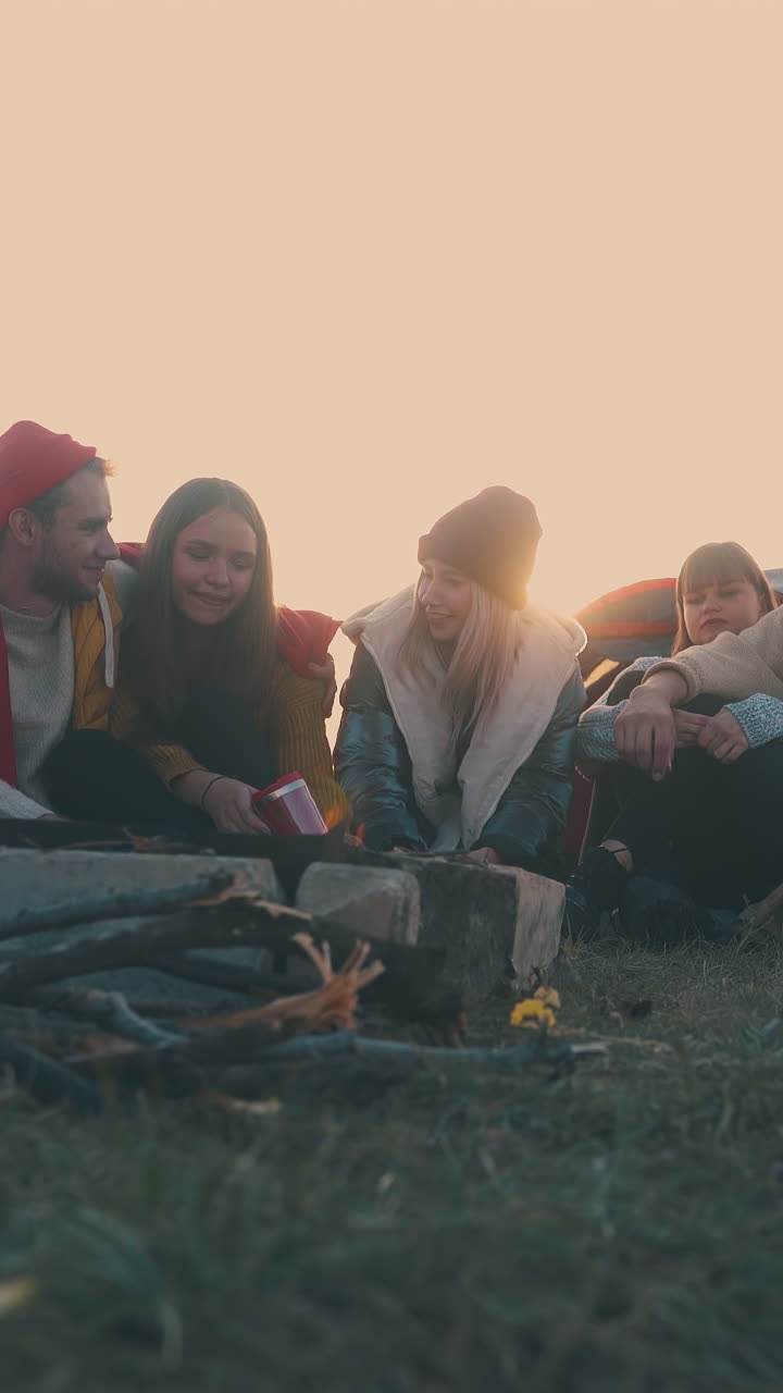 overweight girl takes photo of friends at burning bonfire on hilly river bank at sunset in autumn evening