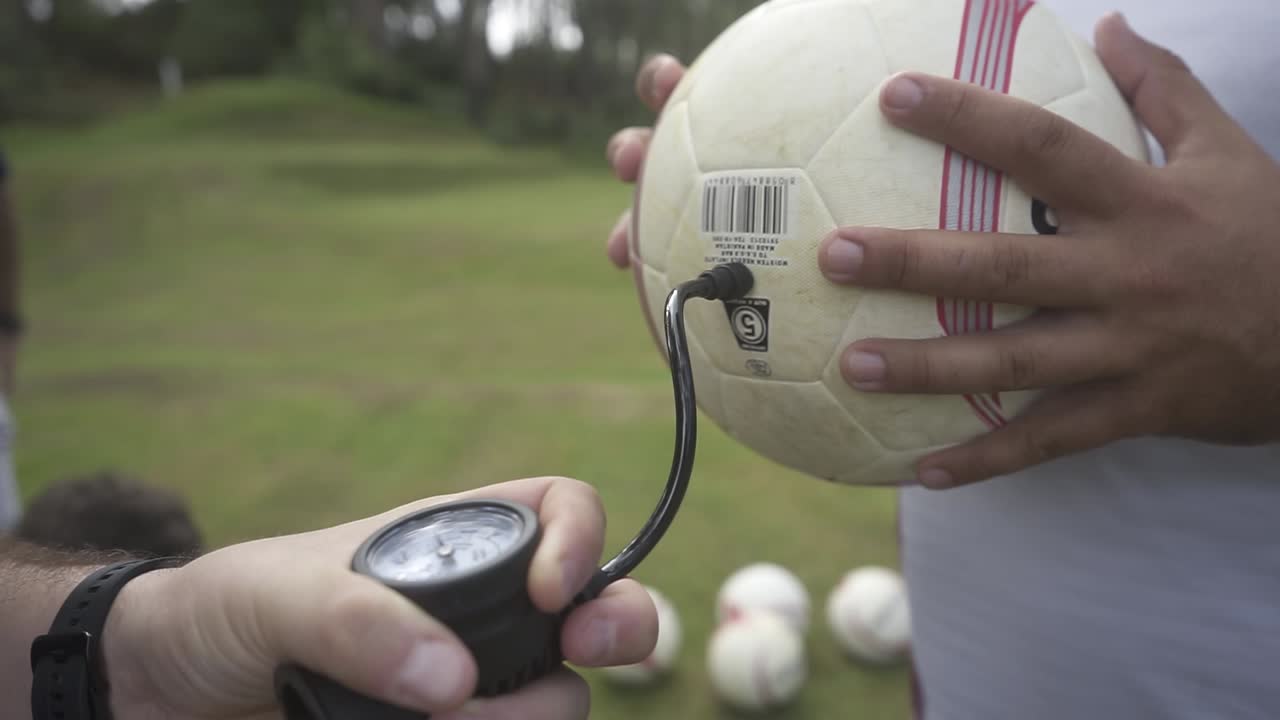 Soccer coach carefully preparing soccer ball, using hand pump to inflate and verify pressure before match on sunlit grassy field