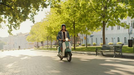 Un-Mensajero-Con-Pelo-Rizado,-Con-Un-Casco-De-Ciclomotor-Blanco-Y-Una-Camisa-Vaquera,-Conduce-Su-Ciclomotor-Verde-Por-Una-Calle-Con-Hermosos-árboles-Verdes-Y-Césped-En-Una-Ciudad-De-Verano.