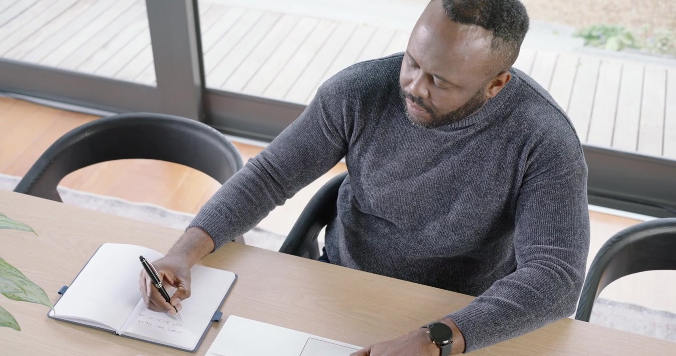 African American man working on project typing laptop adjusting watch writing notes at wooden table