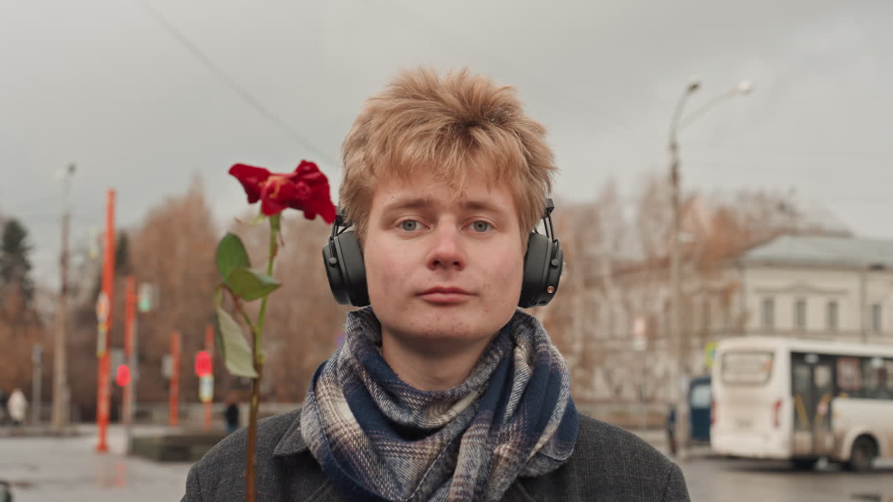 Creativo bianco con le cuffie che tiene una rosa, poeta di strada che offre un fiore e poi fa un gesto offensivo, sfondo di città autunnale, sciarpa e cappotto, sorrisetto giocoso, ritratto urbano spontaneo con cielo nuvoloso e fermata dell'autobus.