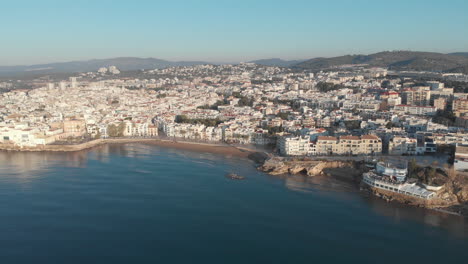 Drone-flies-towards-coastal-town-lit-by-warm-morning-sun-as-gentle-sea-waves-quietly-roll-onto-the-shore