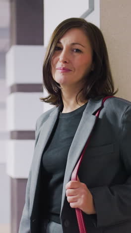 professional businesswoman in gray suit with red handbag standing confidently in office hallway, looking to side with calm expression, modern office background with sleek design