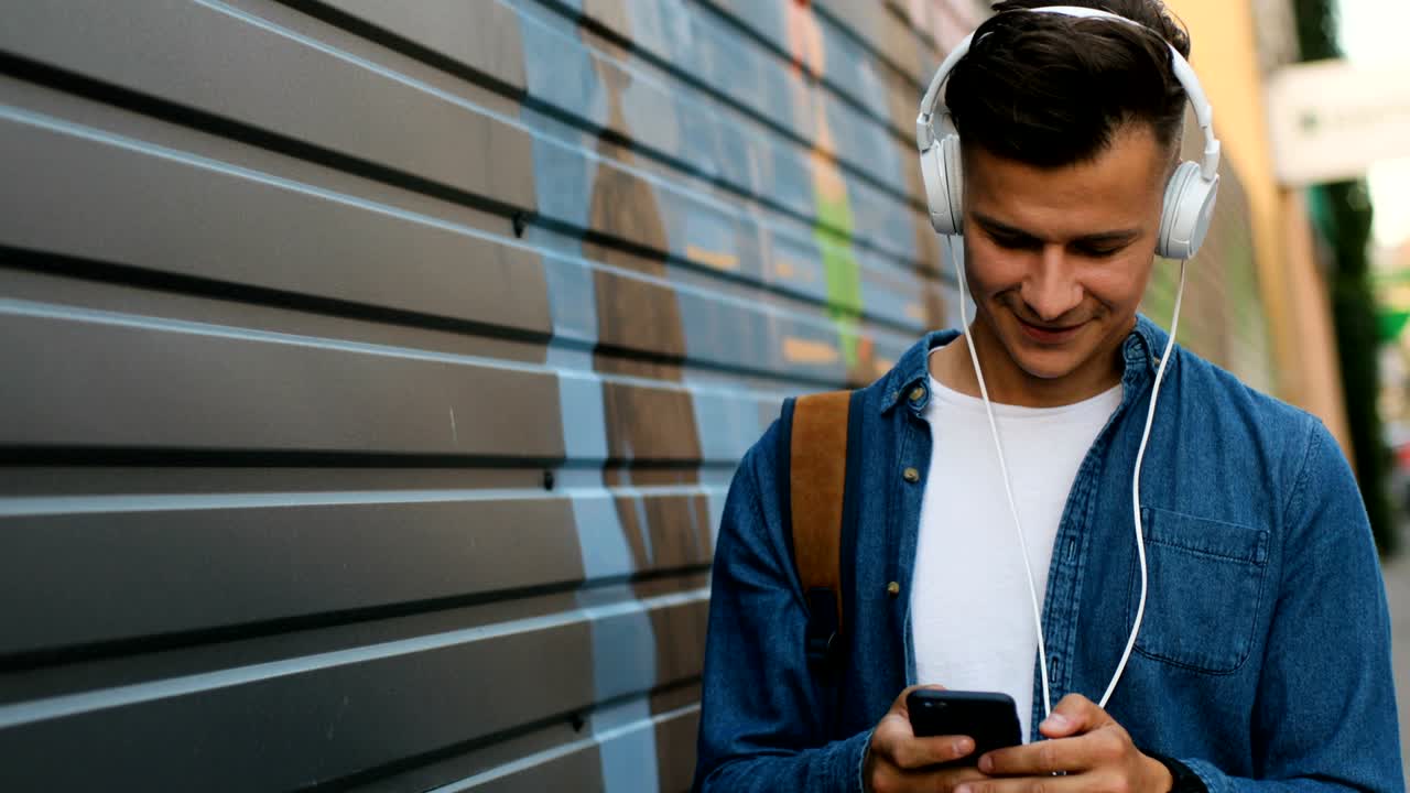 Portrait of young man in the head phones listening the music and using smart phone on the city background.