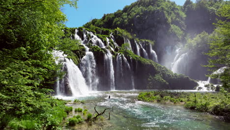 misty waterfalls plunging over lush green cliffs with clear flowing river in foreground. plitvice lakes national park, croatia. drone tilt-up reveal