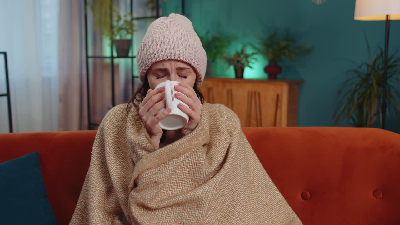 Sick young woman wear hat wrapped in plaid sit alone shivering from cold on sofa drinking hot tea