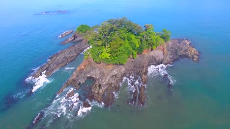 A-stationary-aerial-shot-of-a-tiny-rocky-island-with-a-small-whitewash-waves-splashing-against-the-shore-the-coast-and-bouncing-back-to-sea