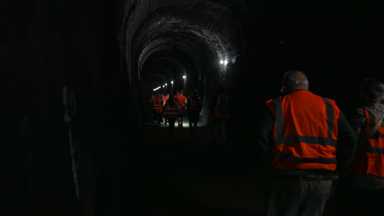 A group of people wearing high-visibility vests walking through a dark tunnel underground. The tunnel's brick structure and low lighting create a sense of exploration or industrial inspection