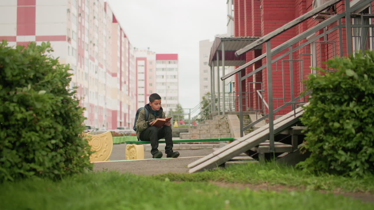 Ребенок сидит на улице и увлеченно читает книгу, перелистывая страницы. За плечами рюкзак, рядом колышется зелень, вдалеке — здание из красного кирпича