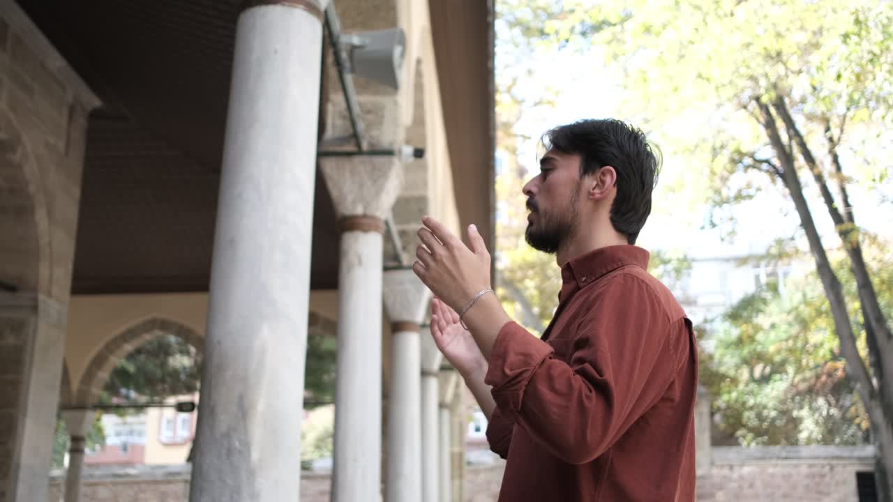 Side view of a young bearded man praying with open hands in the garden of the mosque, people who believe in Islam pray to god
