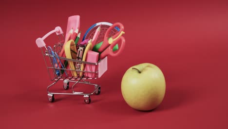 Close-up-of-shopping-trolley-with-school-items-and-apple-on-red-background