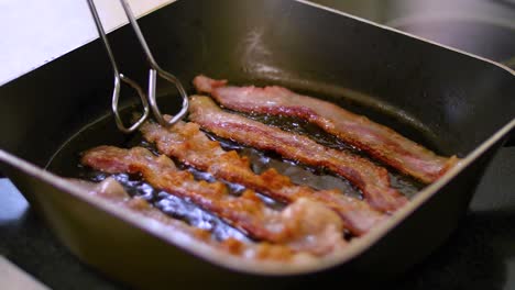 Bacon-strips-sizzling-in-frying-pan-are-flipped-by-tongs,-closeup