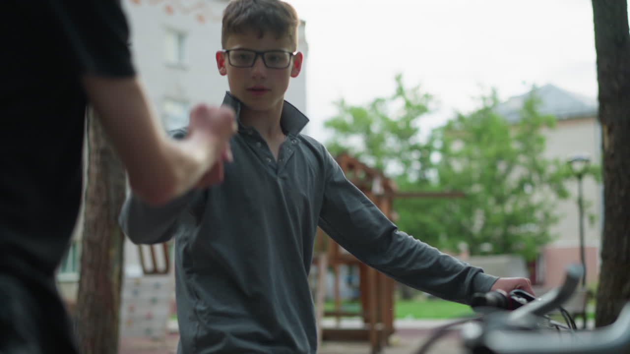 자전거를 타고 다니는 두 형제는 장난스럽게 상호 작용합니다. 회색 을 입은 한 형제는 다른 형제의 손목을 기 위해 손을 습니다. 둘 다 미소 짓고, 배경에는 놀이터와 주거 건물이 있습니다.