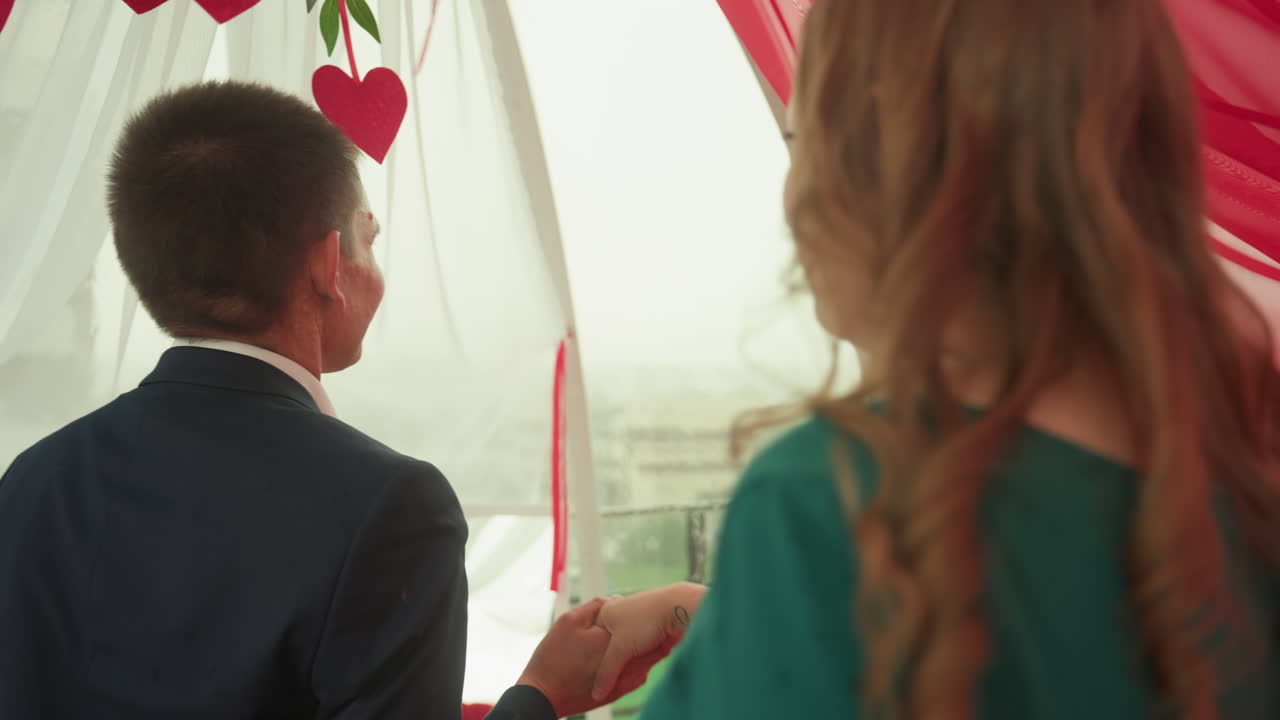 Romantisk date på taket, et par ser utover horisonten. Mann i dress og kvinne i grønn kjole sitter ved et bord med levende lys under flytende røde draperinger, tulipanbukett og frokostfat, de smiler og peker.