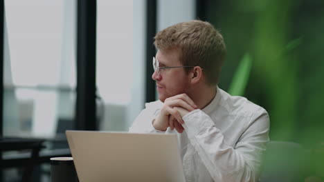 Focused-concentrated-young-businessman-sit-at-desk-look-at-laptop.-Authentic-portrait-of-young-confident-businessman-looking-at-camera-with-laptop-in-office