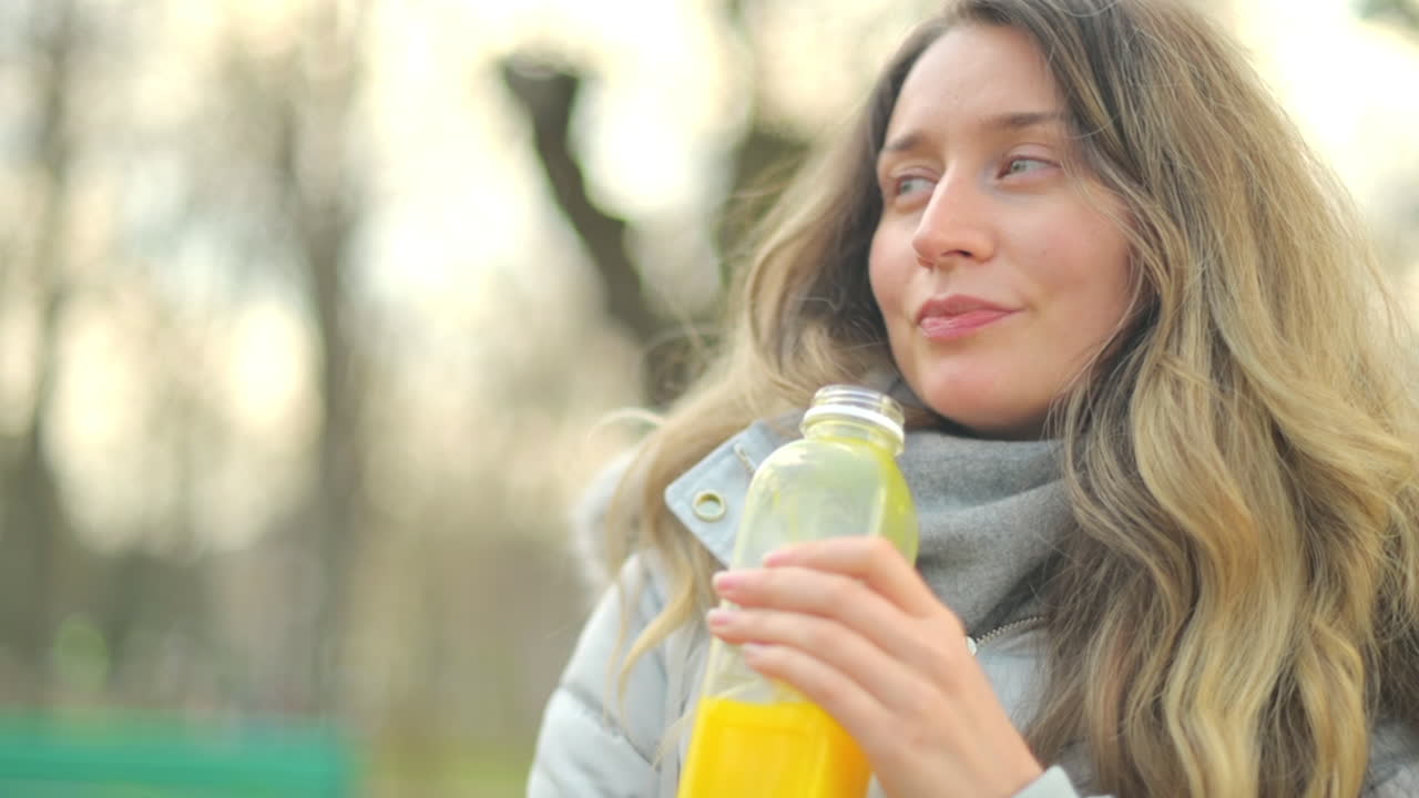 Woman in a puffer jacket drinking orange juice in a park