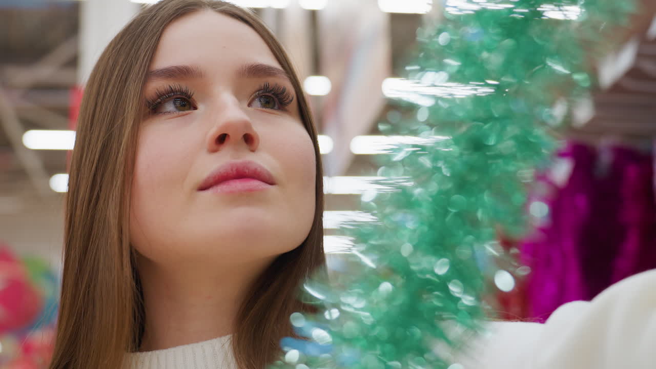 Woman with warm expression inspecting green shiny tinsel length in store with bokeh light effect, carefully evaluating decoration options for festive holiday shopping, surrounded by colorful decor