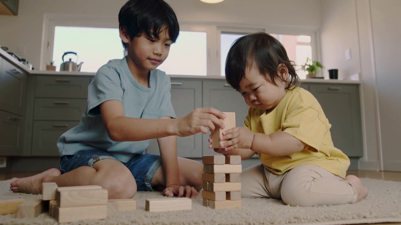 Selecting blue-shirted child starting block tower in kitchen, guiding toddler placing wooden blocks