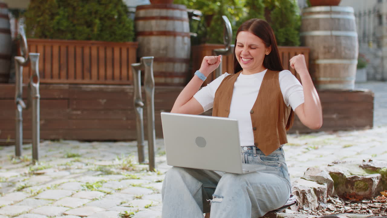 una joven feliz que trabaja en una computadora portátil gritando de alegría levanta las manos en la calle de la ciudad