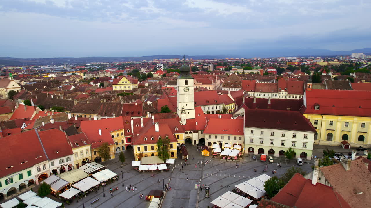 Lotniczy widok z drona na Stare Miasto w Sybinie o zachodzie słońca, Rumunia. Wielki Rynek z Wieżą Rady i starymi budynkami wokół, wąskie uliczki, ludzie, oświetlenie