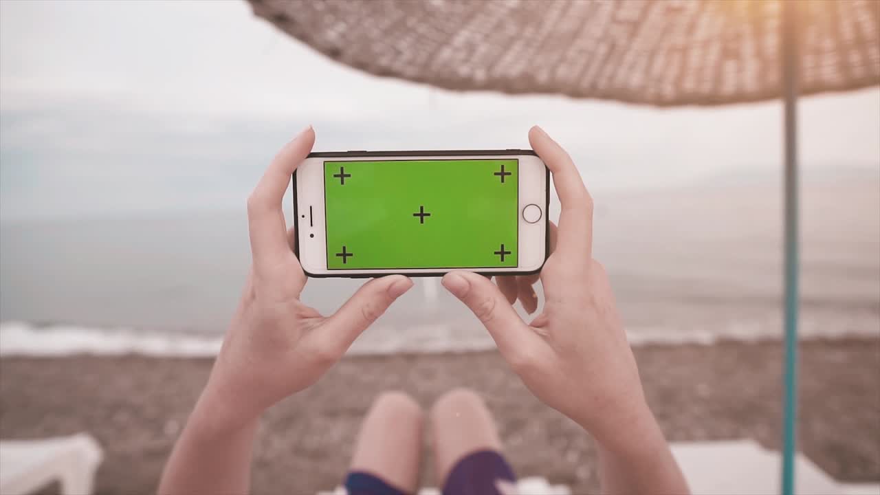 mujer de vacaciones en una cama de sol, cerca del océano y teléfono móvil vertical con pantalla verde, canal alfa y marcas de seguimiento. maquillaje de video de teléfono inteligente y concepto de verano.