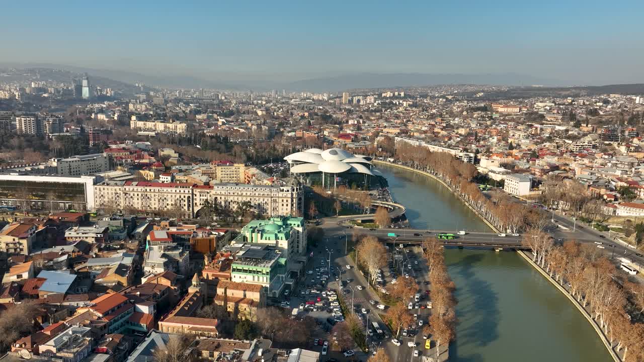 Aerial panorama of the Kura River flowing through central Tbilisi, surrounded by modern buildings, bridges, and riverfront streets under clear weather