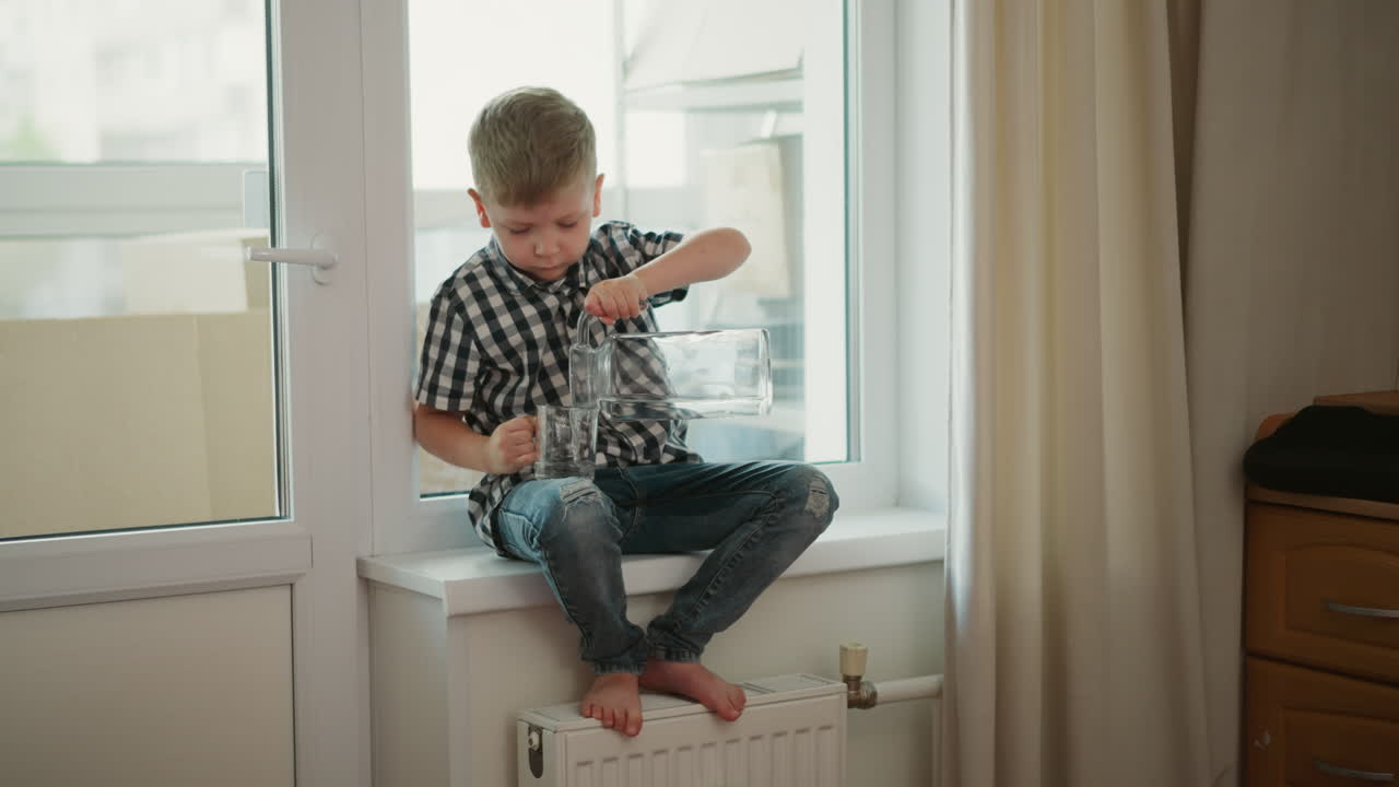 Focused kid wearing plaid shirt sitting on windowsill barefoot pouring water from glass jar in right hand into clear cup in left hand lifting cup to drink inside