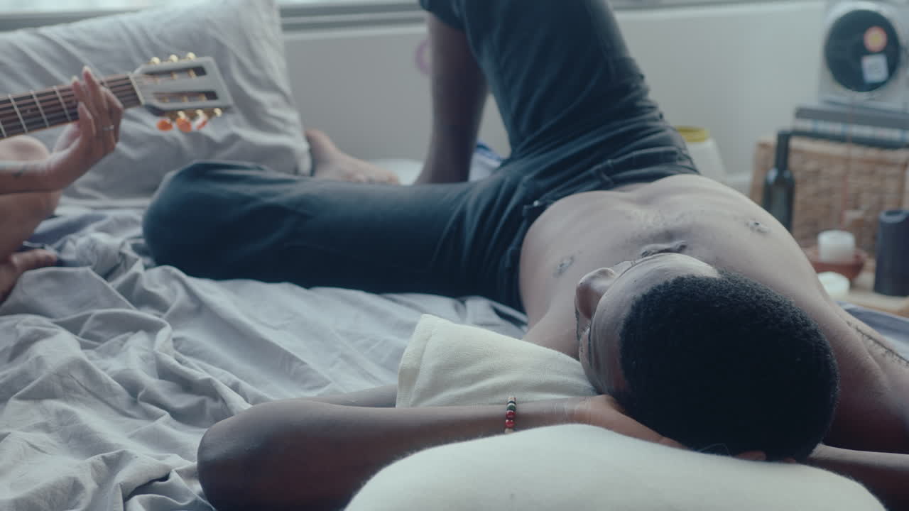 Girl Playing the Guitar and Singing for Her Boyfriend Resting on Bed