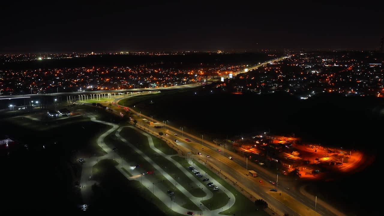 Drone video showcases Asuncion's Costanera and Heroes of Chaco Bridge. Night skyline glows with city lights below. Captures expansive urban landscape from above