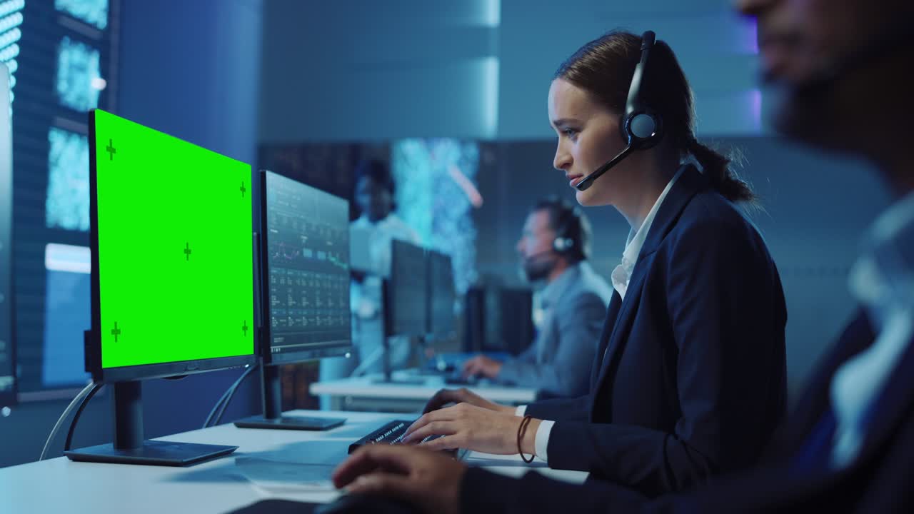 un especialista de soporte técnico de clientes feliz y confiado teniendo una llamada de auricular mientras trabaja en una computadora con pantalla verde en una sala de monitoreo llena de colegas y pantallas de visualización.