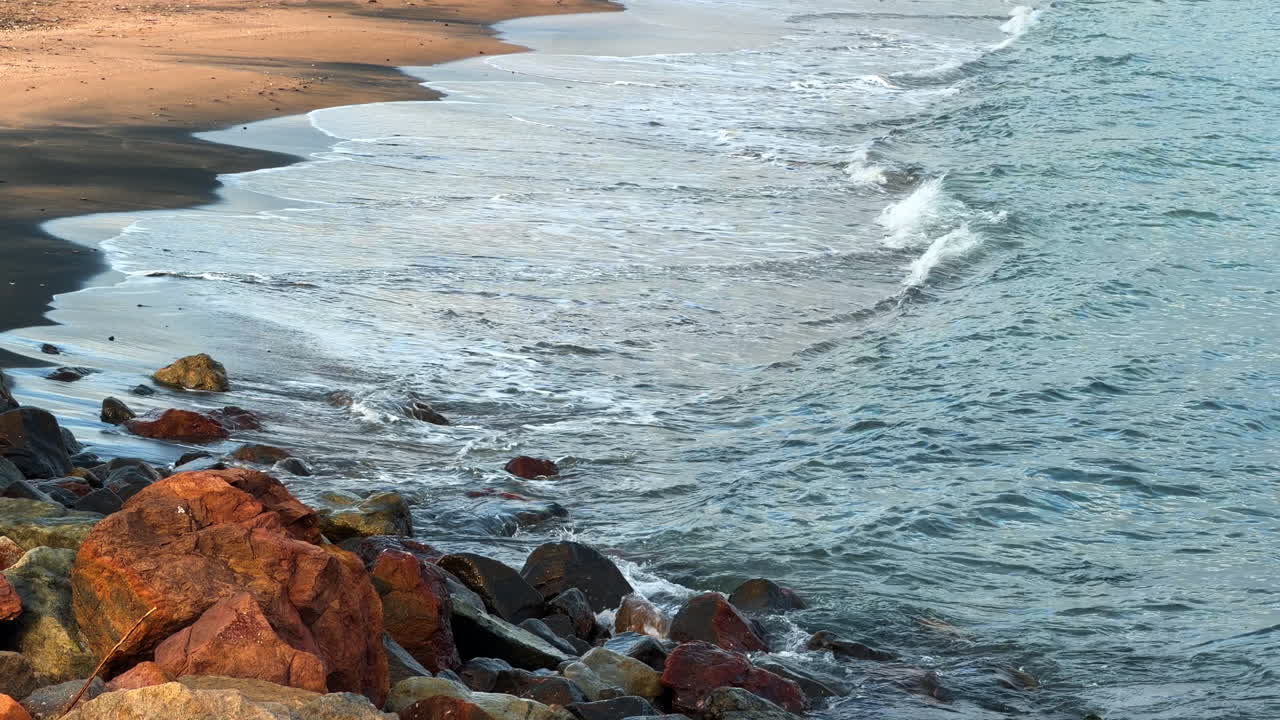 Spiaggia costiera con sabbia scura e grandi rocce rosse. Litorale caratterizzato da sabbia scura bagnata, grandi pietre bruno-rossastre e piccole onde oceaniche che raggiungono la riva