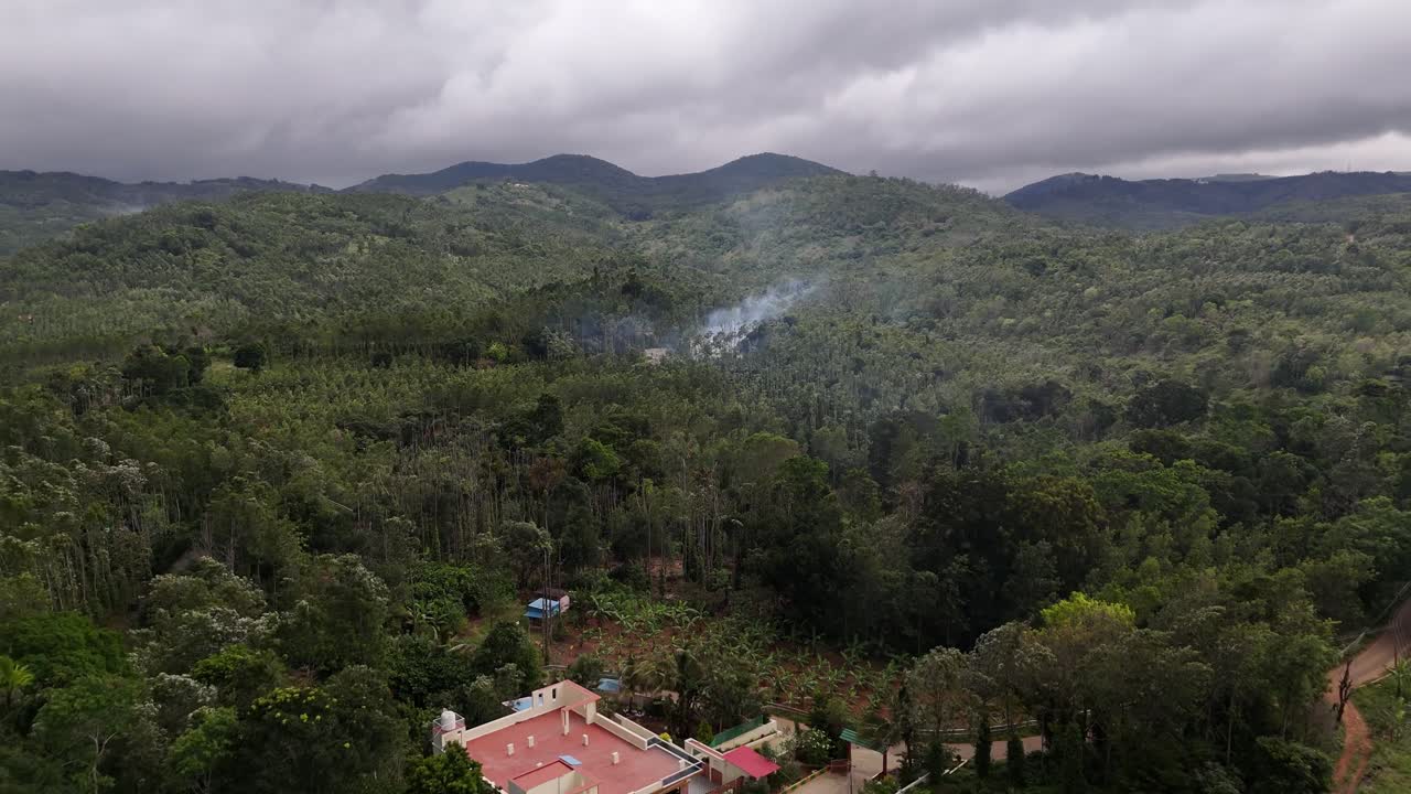 Moody aerial shot of dense, green hillside forests under a cloudy, overcast sky. Smoke rises mysteriously from a hidden clearing, suggesting environmental activity or a campfire