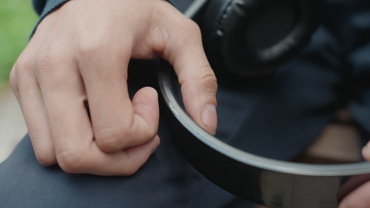 yakın çekimde kulaklık baş bandını tutan el, gergin bir öğrenci bankta oturmuş parmak uçlarıyla bandı takip ediyor, deri kulaklık kabı görülebiliyor, kısık dış mekan tonları, günlük ceket ve kot pantolon, dokunsal detay ve sığ alan derinliği