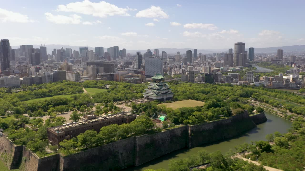 Wide aerial flying towards historic Osaka Castle with park, moat, skyscraper, and city in Osaka, Japan