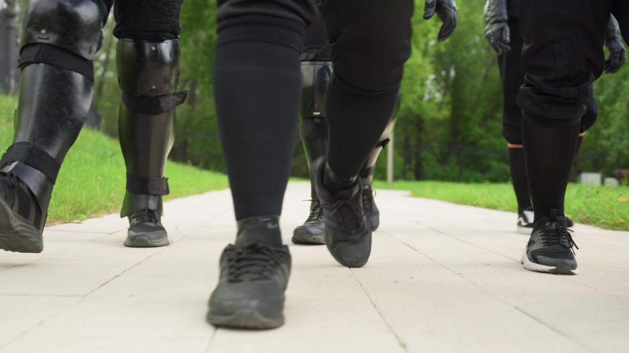 Close up of multiple people walking in unison wearing black clothing and protective leg gear on paved path with green grass and trees in background. synchronized movement highlights their