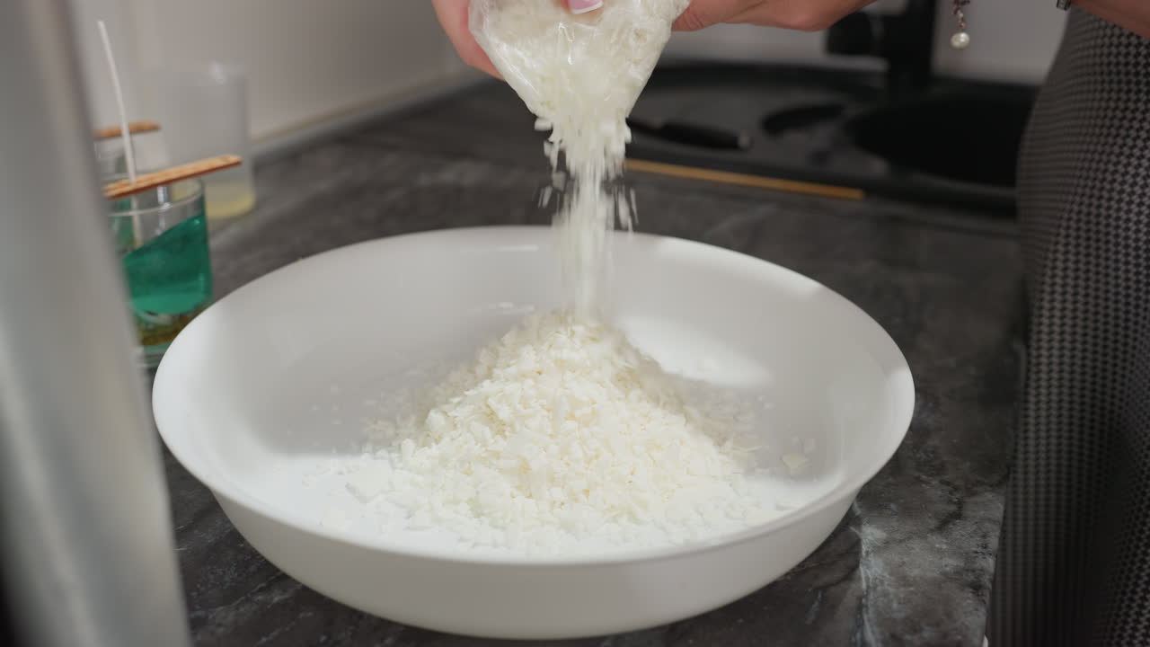 hand view of fair skinned woman pouring unmelted wax into large white bowl on kitchen counter with candle containers in background showing wax sticks set in cups during homemade candle preparation