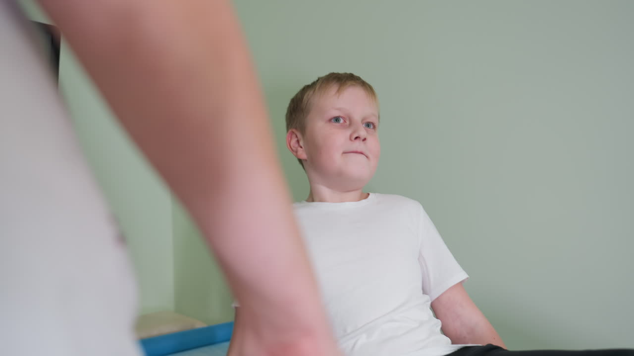Sick young boy sits on medical bed preparing for examination by female doctor showing nervousness as he sticks fingernail in trouser pocket while awaiting diagnostic care in clinical environment