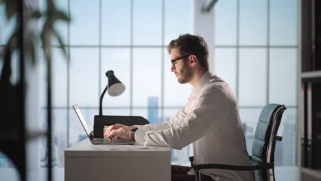 portrait of thoughtful successful businessman working on laptop computer in his big city office. digital entrepreneur does data analysis for e-commerce strategy.