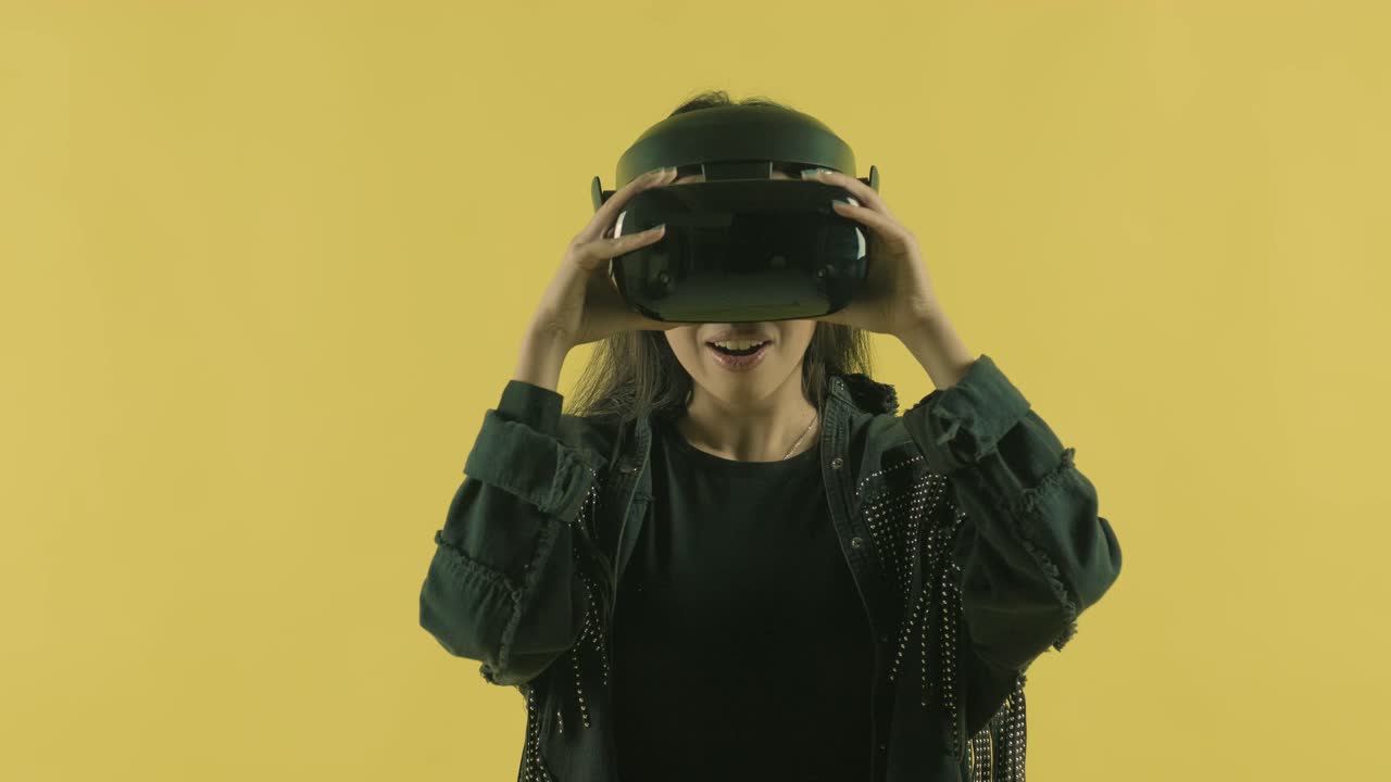 Young woman puts on vr headset, looks around and wonders how amazing  in the VR world. Virtual reality helmet on the yellow background