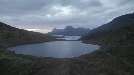 Vista-Aérea-De-Drones-A-Baja-Altura-Sobre-Un-Lago-Hacia-Las-Montañas,-Amanecer-En-Lofoten-Nublado,-Noruega