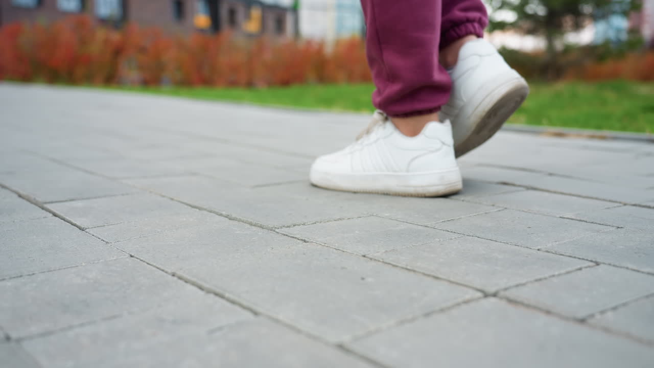 Leg view of female jogger striding on grey tiled pavement amid urban park with budding spring foliage and bars in background conveying active lifestyle momentum with white sneakers