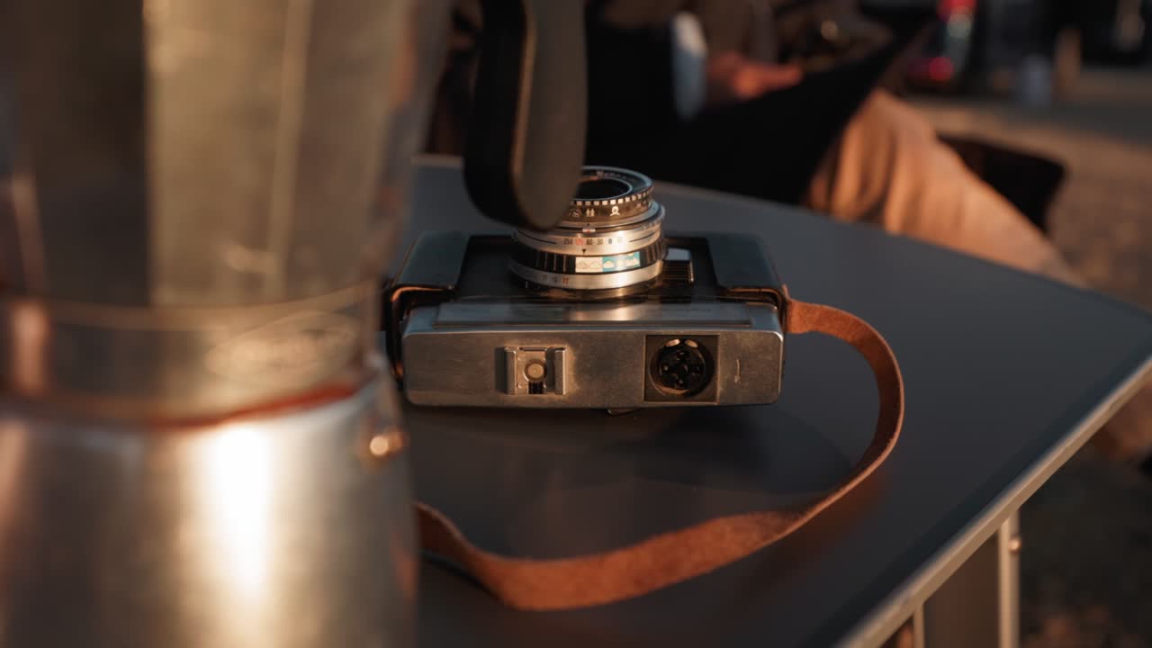 Close-up on vintage camera with leather strap and moka pot, warm tones and human presence in cozy indoor setting.