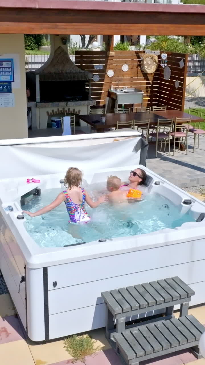 Vertical drone shot circling mom and kids relaxing in a jacuzzi, sunny day